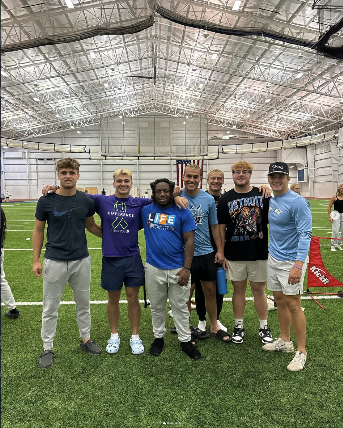 Seven young men stand together on an indoor sports field, posing for a group photo. An American flag hangs in the background, highlighting their unity as members of the Fellowship of Christian Athletes (FCA).